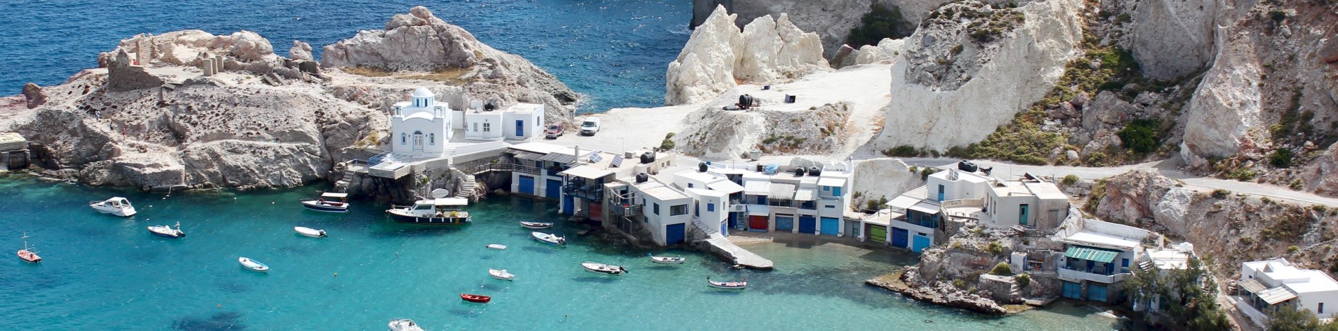White buildings and jagged rocks on the coast of a Cyclades island surrounded by blue water with boats