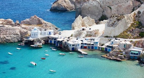 White buildings and jagged rocks on the coast of a Cyclades island surrounded by blue water with boats (1)