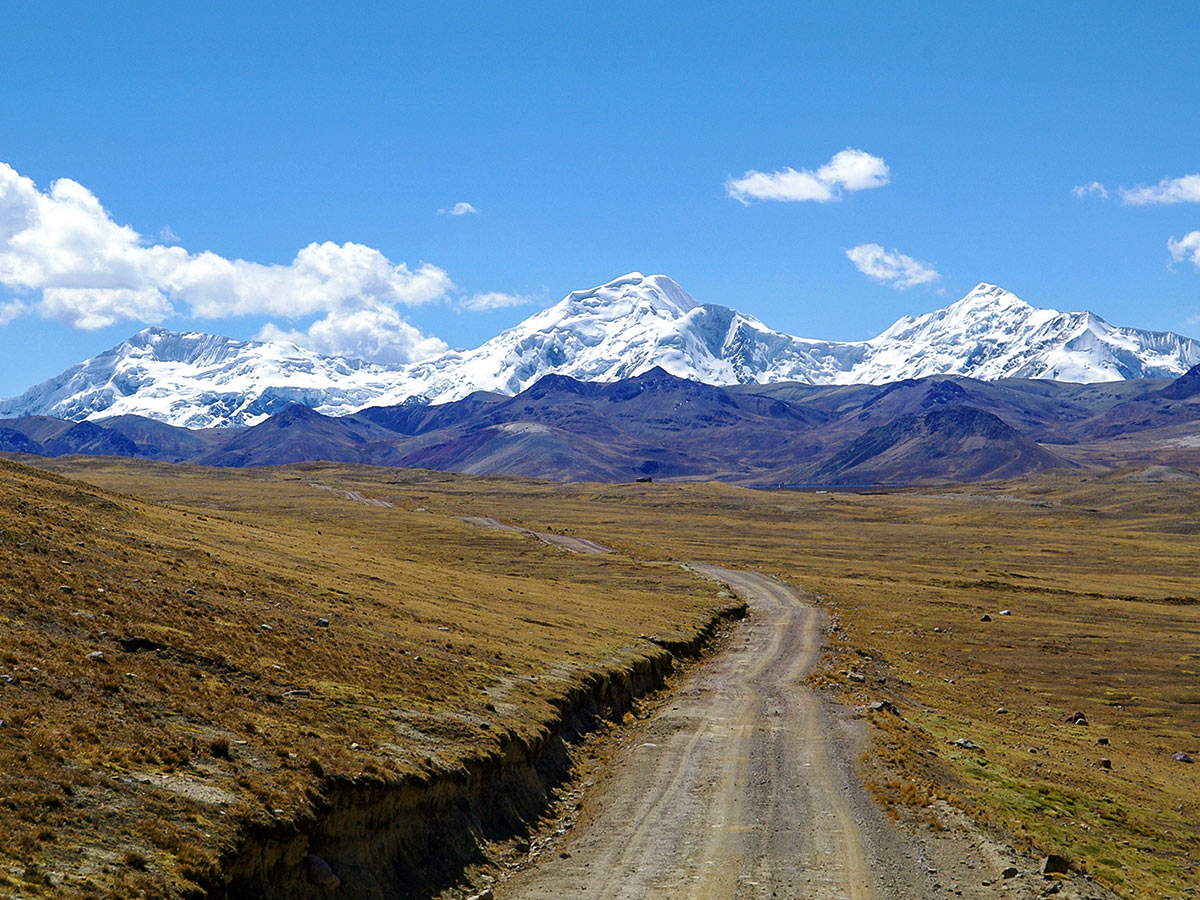 A dirt road cutting through an open plain with two layers of snow-covered mountains in the background