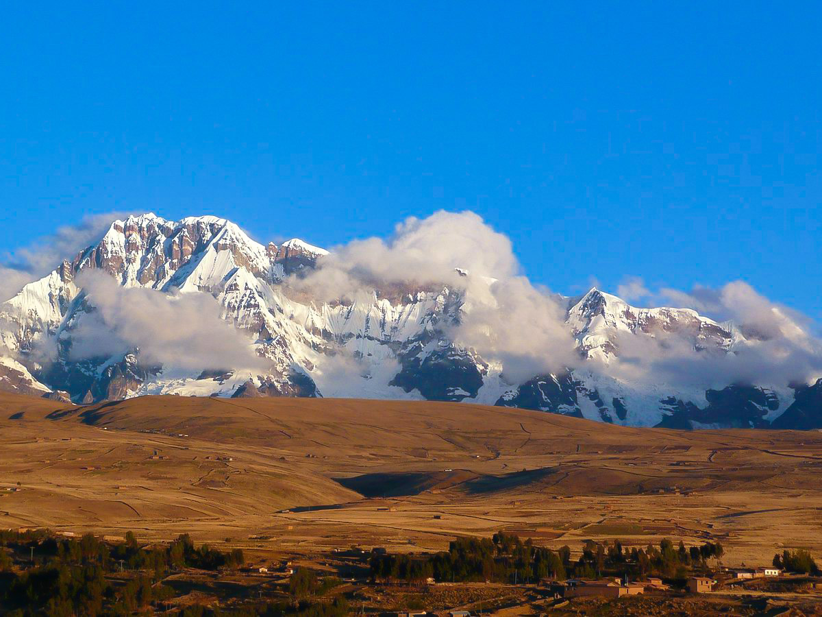 A small village on red-tinted hills with tall, snow and cloud-covered mountains in the distance