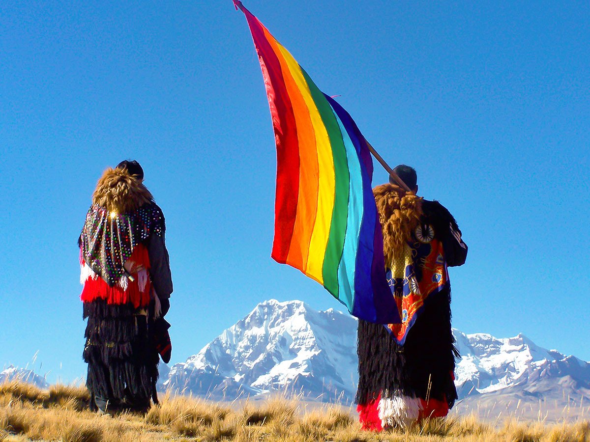 Traditionally dressed indigenous men with a rainbow flag looking out over mountains