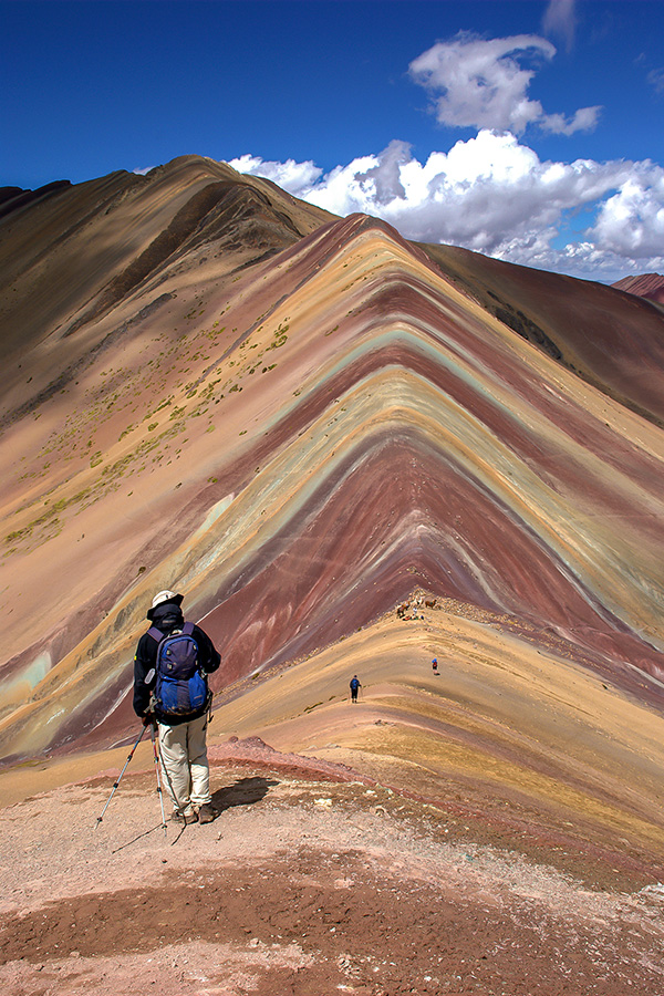 A person with a backpack and trekking poles walking along a striped, multicoloured ridge of rock in Peru