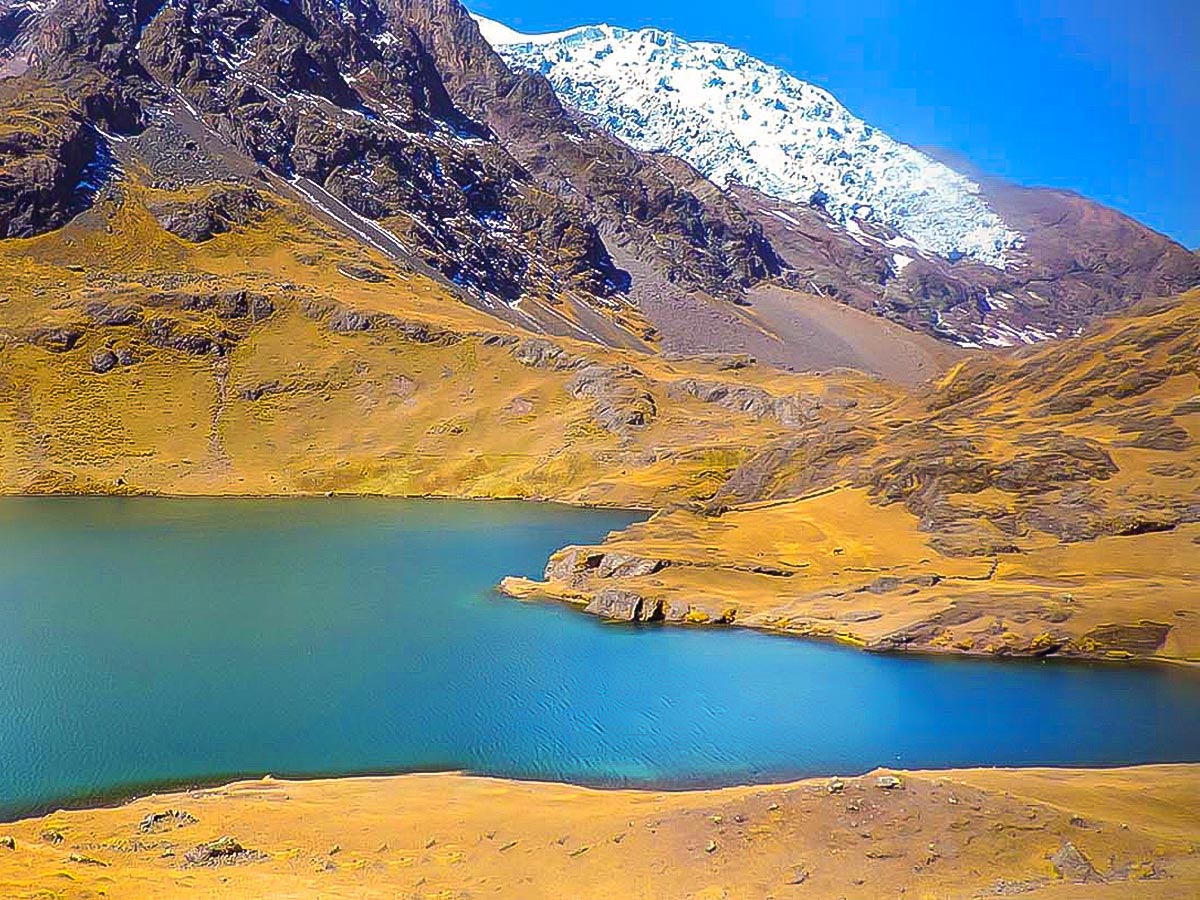 A bright blue lake surrounded by yellow hills and a snow-covered mountain in the distance